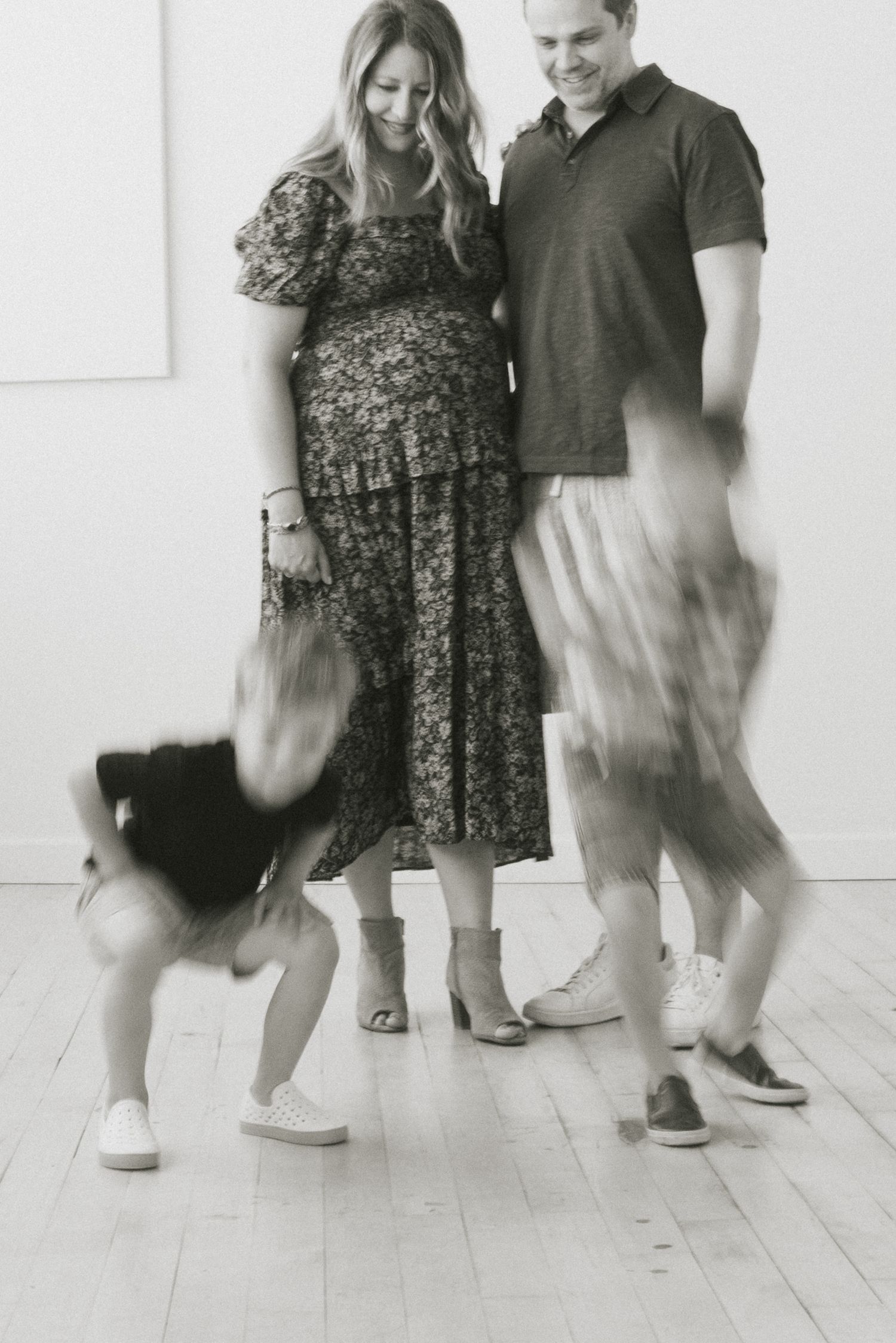 Black and white family photo showing blurred motion of children running while couple stands together on wooden floor.