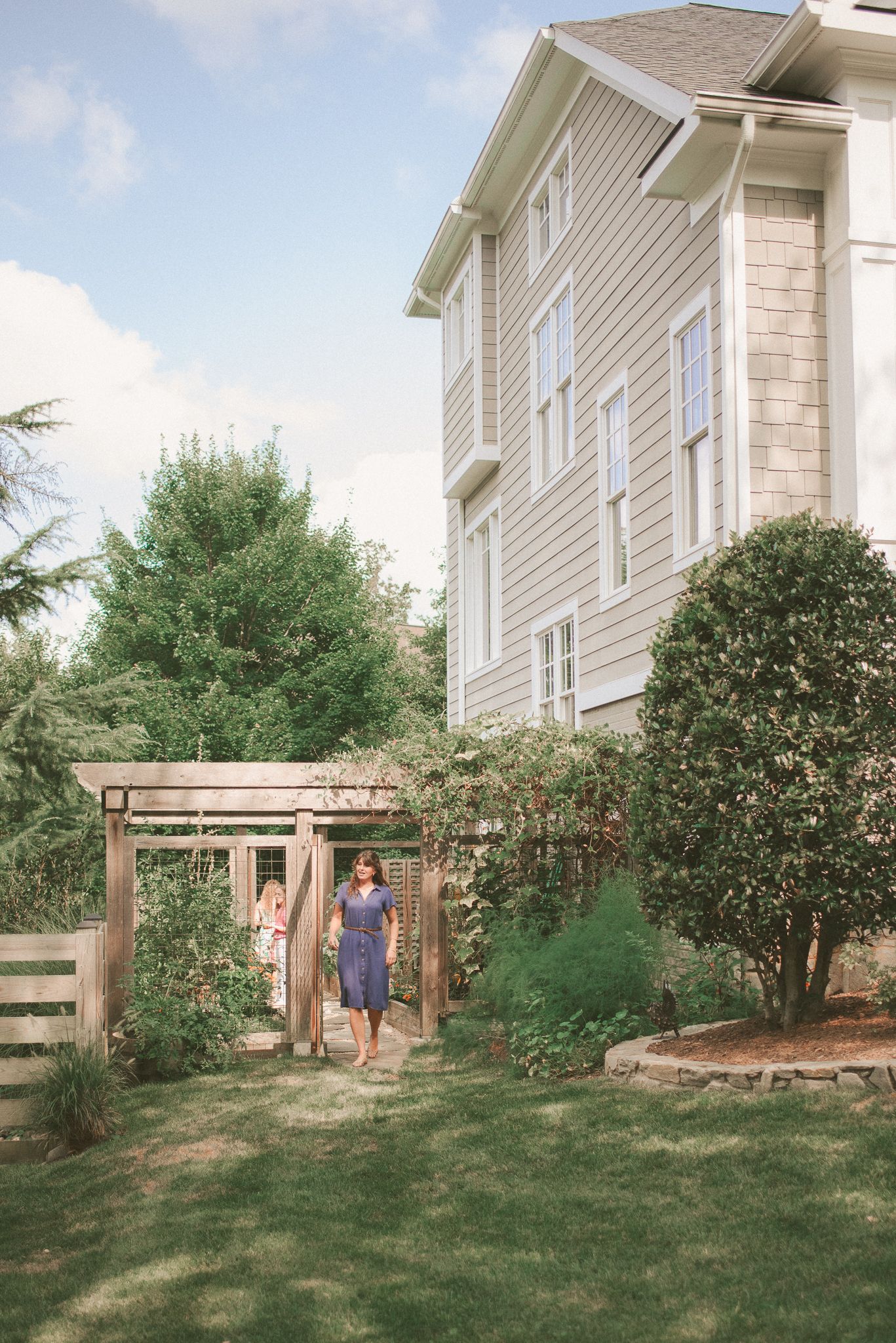 A wooden garden arbor stands in a backyard against the backdrop of a white house with green landscaping.