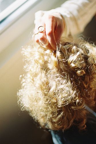 A hand with a gold ring gently holds a curl of voluminous blonde curly hair near a sunlit window.