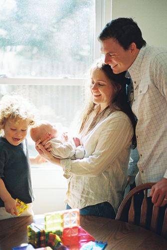 Family of four gathered indoors, parents smiling at newborn baby while young toddler plays with colorful blocks on table.