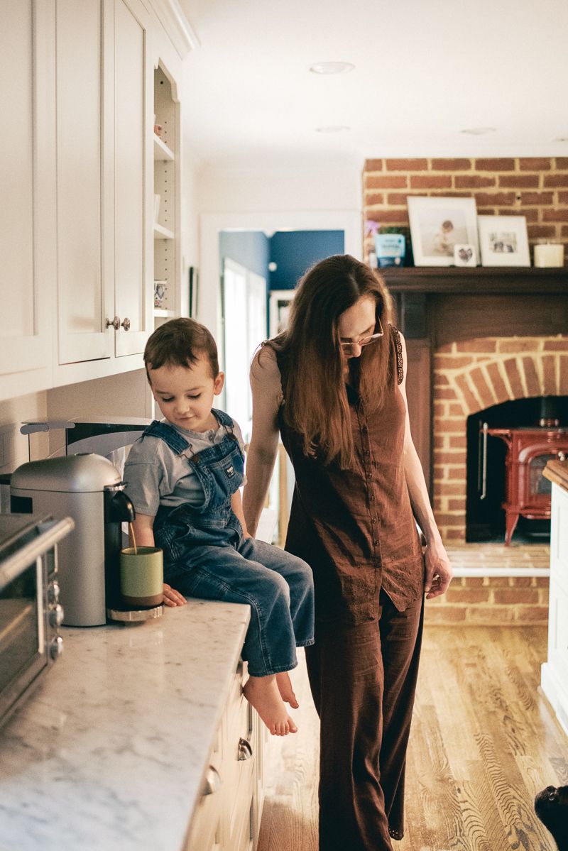 Mother in brown jumpsuit watching toddler boy sitting on kitchen counter near fireplace.