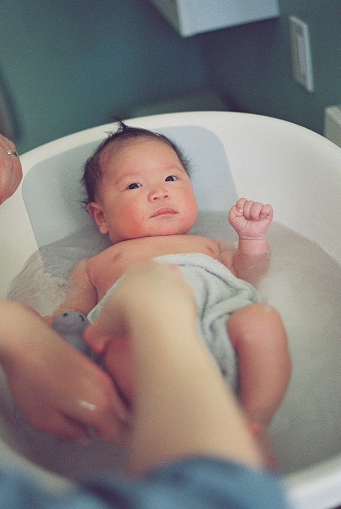 Newborn baby with dark hair relaxes in a small white infant bathtub during first bath.