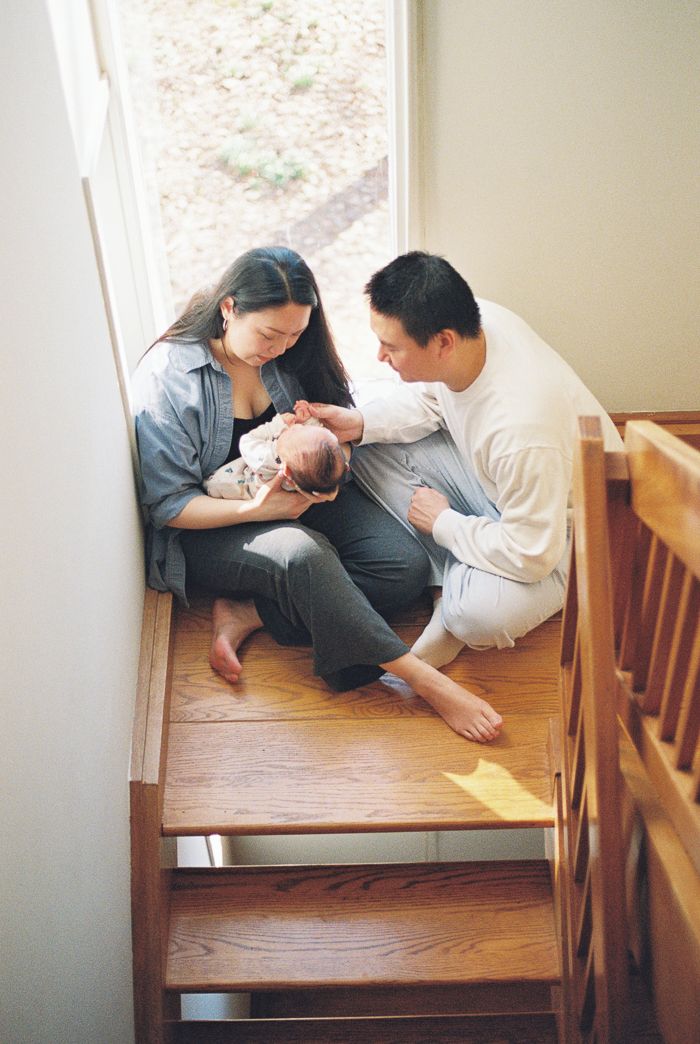 A couple sits on wooden stairs cradling their newborn baby, sharing a tender family moment near a sunlit window.