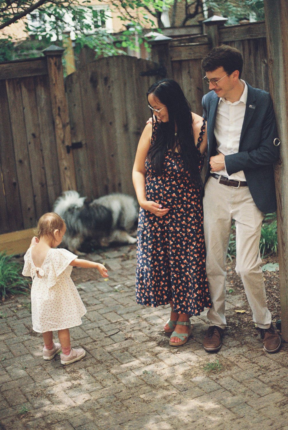 A smiling couple stands arm in arm on a brick patio beside a wooden fence as a toddler in a white dress and a fluffy dog look on.