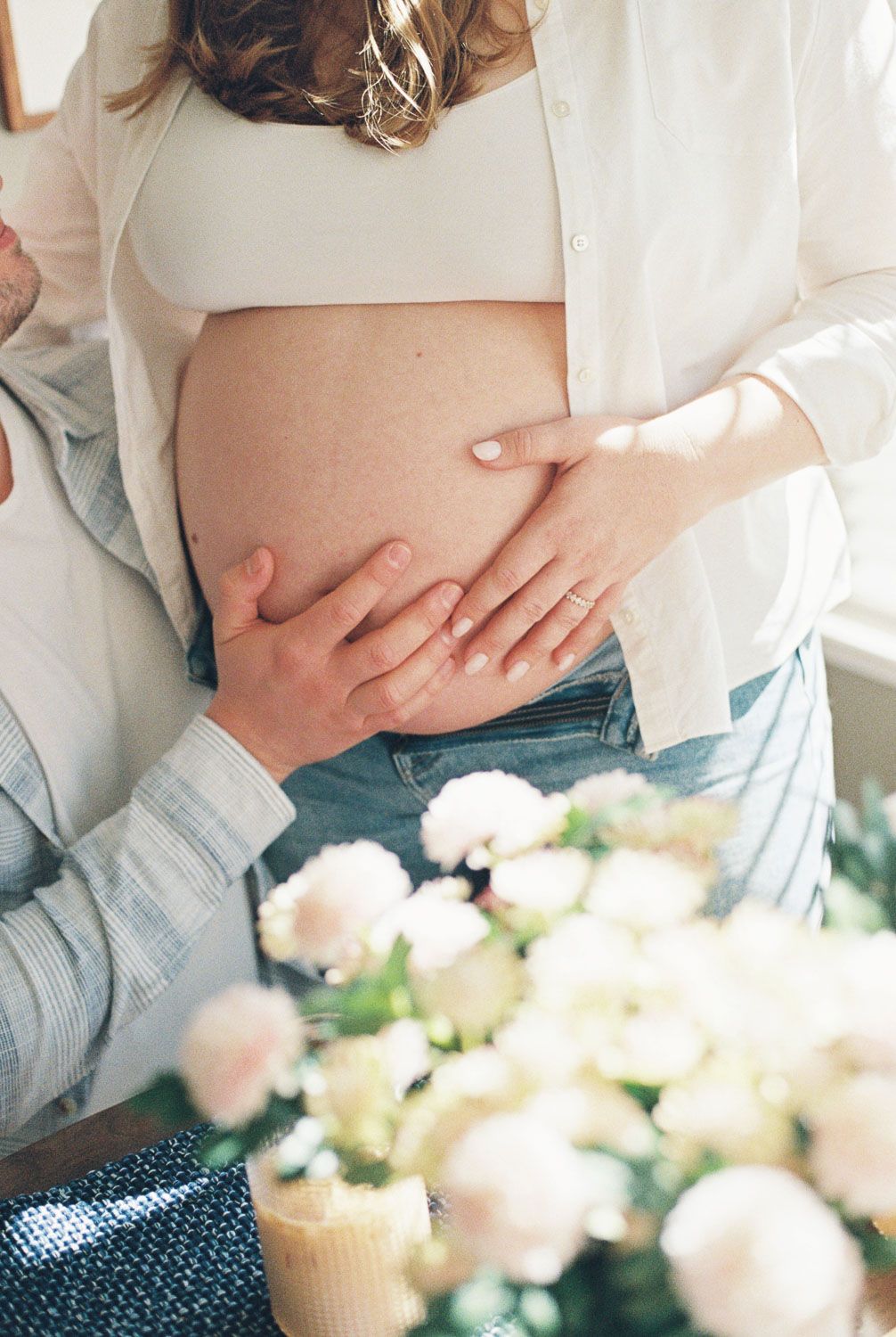 Pregnant woman in white shirt and jeans with partner's hands gently cradling baby bump near soft pink flowers.