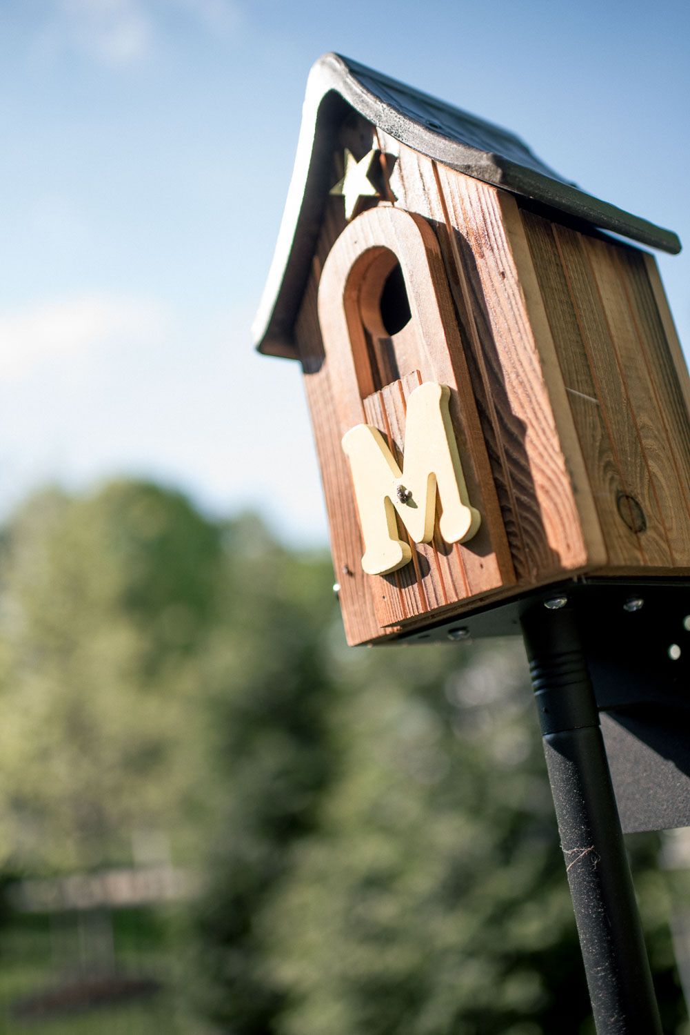 Wooden birdhouse with gold letter M and star decorations mounted on a black pole outdoors.