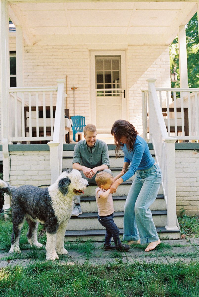 Family with toddler and fluffy sheepdog on front porch steps of white brick home.