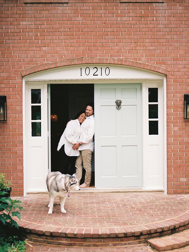 Husky dog stands on brick porch in front of white door with number 10210 while couple embraces in doorway.