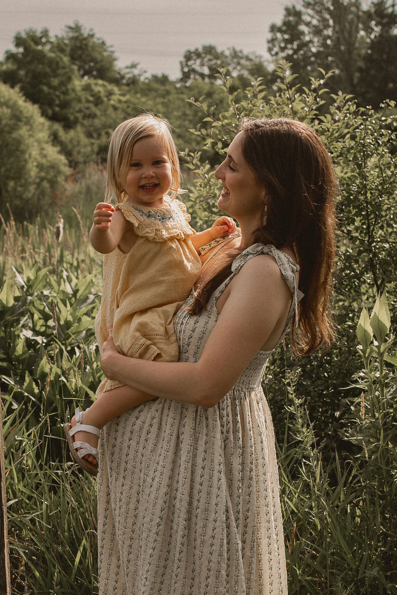 A heartwarming outdoor portrait of mother and daughter in matching light dresses amid lush summer greenery.