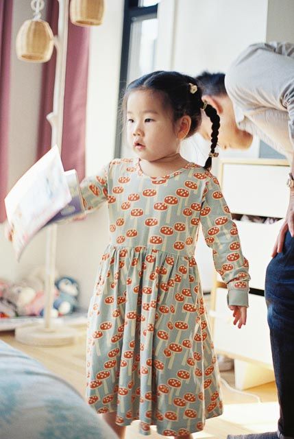 Young girl in a mushroom-print long-sleeve dress with braided pigtails standing in a bright room.