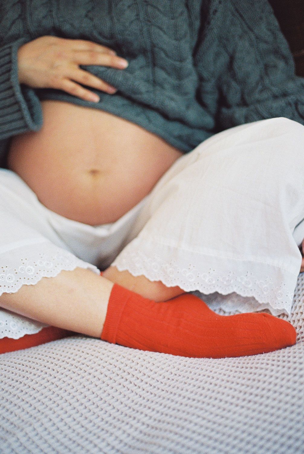 Pregnant woman in green sweater and white skirt with red socks cradling her bare bump.