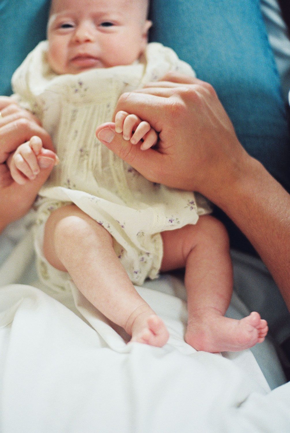 Newborn baby in white onesie grasping an adult's finger while lying on a white blanket.
