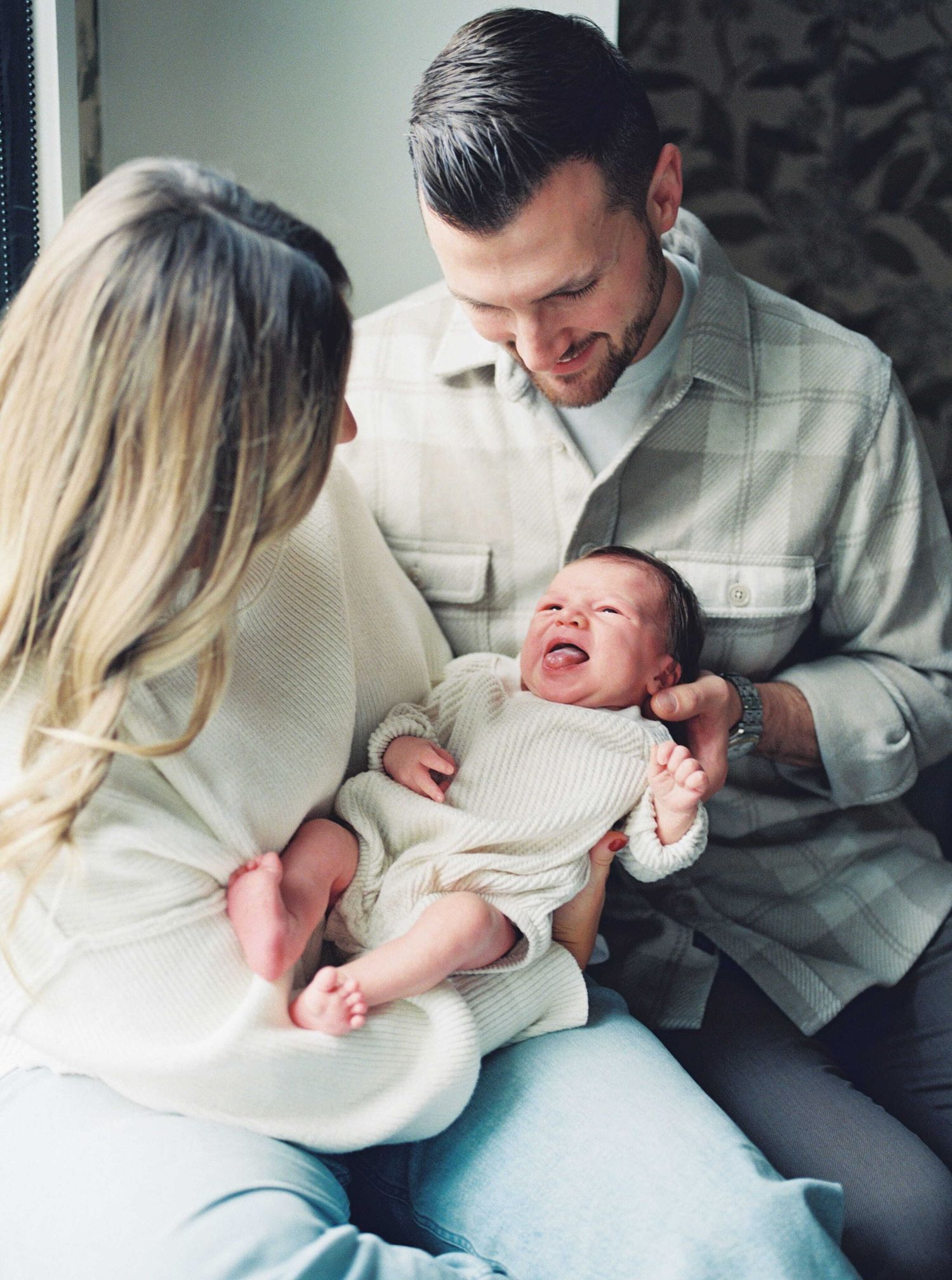 Parents in matching white outfits share tender moments with their baby on a couch during an intimate photo session.