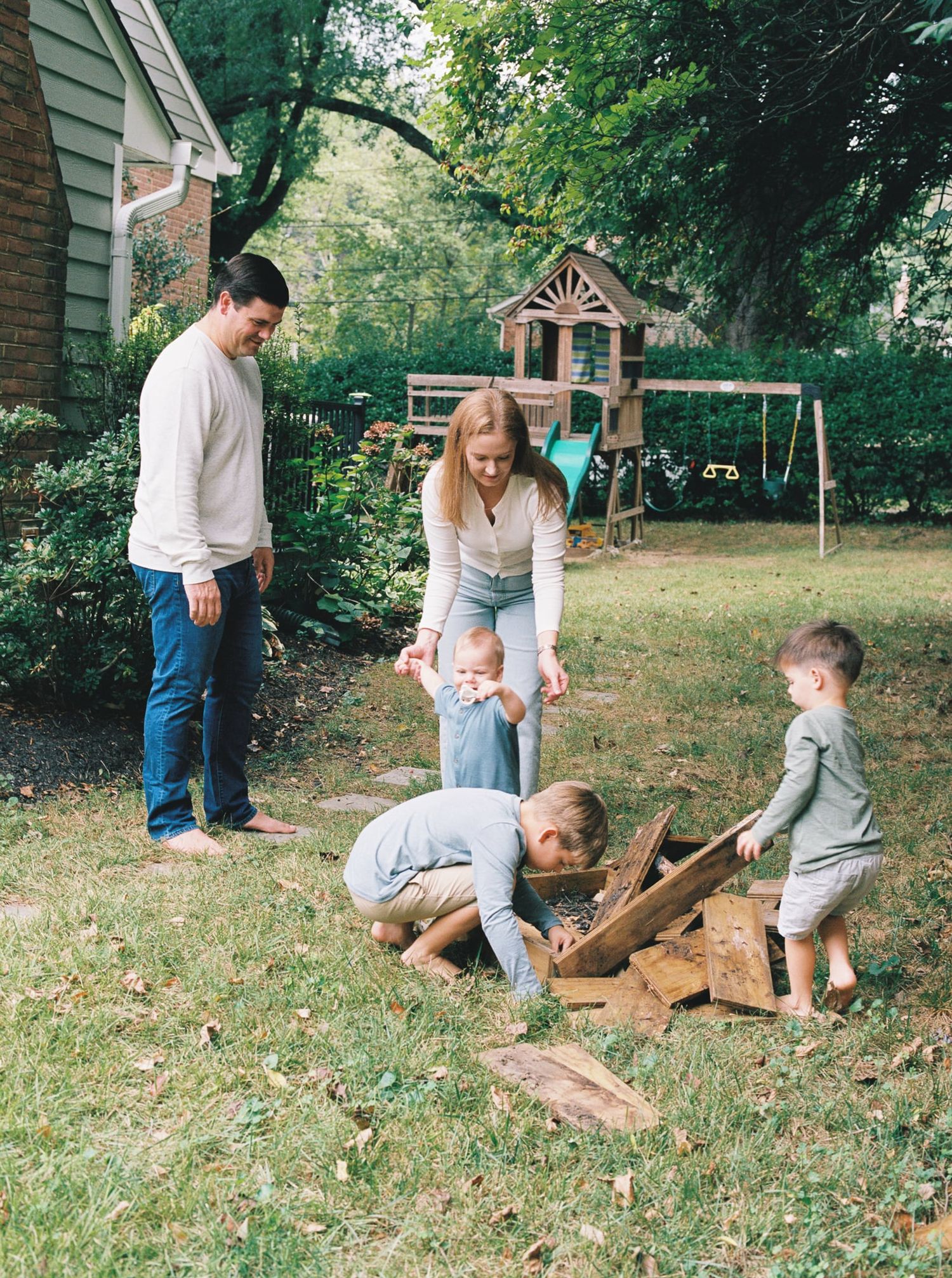 A group gathers around chopped firewood in a backyard with a wooden playhouse and swing set visible in the background.