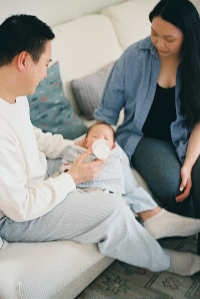 Parents sitting on a couch feeding their newborn baby with a bottle at home.