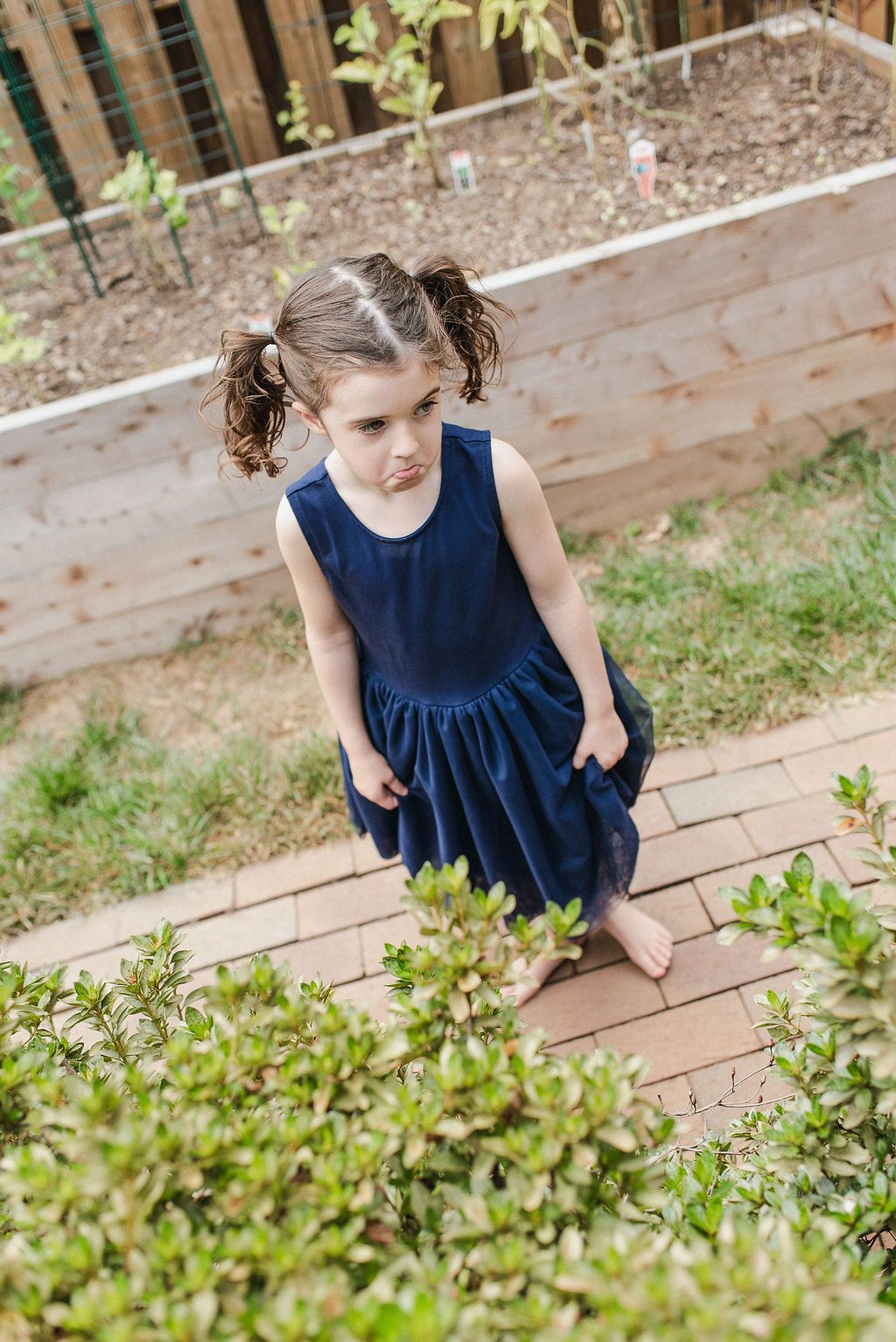 A young girl in a navy blue dress sits on wooden steps in a garden setting.