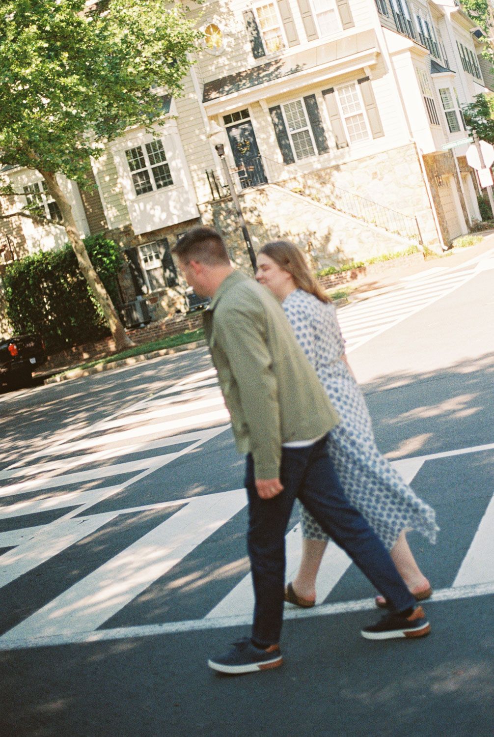 Couple holding hands while crossing a zebra crossing on a sunny street, man in green jacket and woman in blue patterned dress.