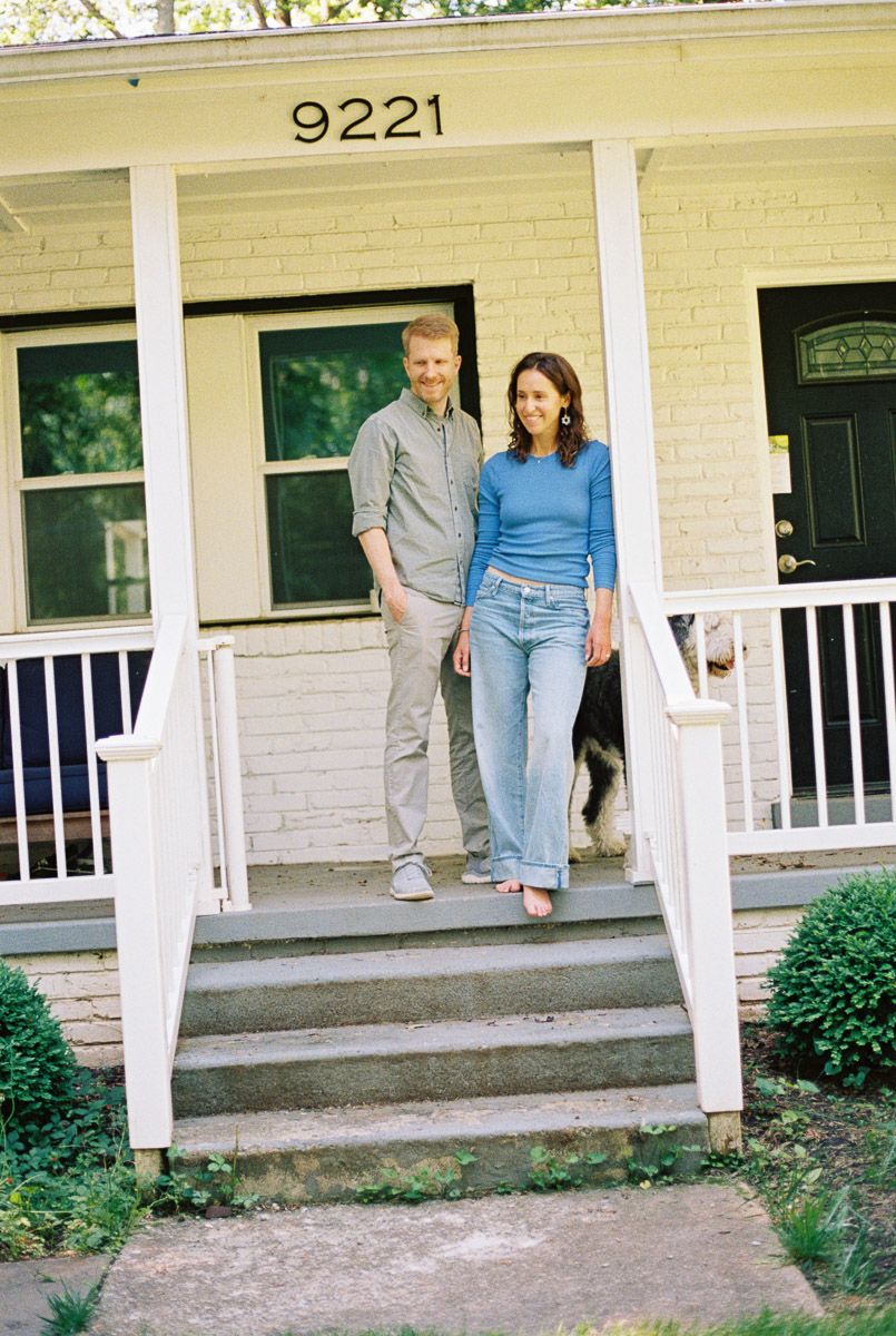 Couple standing on front porch steps of white brick house numbered 9221 with dog visible behind them.