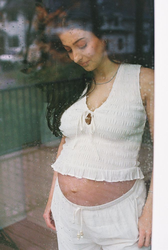 A pregnant woman in a white smocked tank top and drawstring pants stands by a window with a dreamy expression.