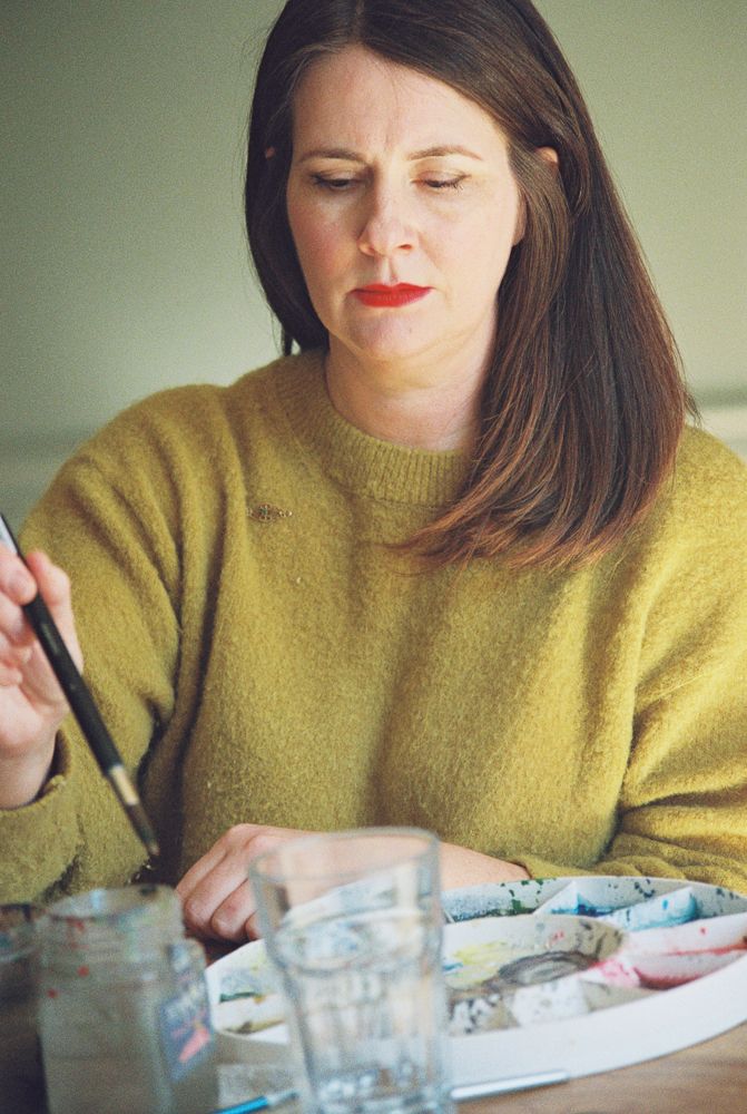 A person wearing a yellow sweater works on detailed artwork at a desk with art supplies.