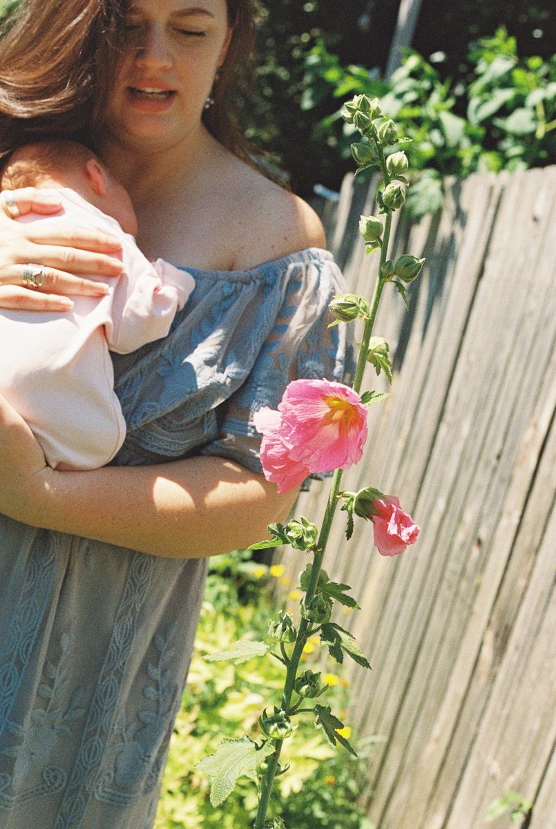 A woman in a blue off-shoulder dress cradles a sleeping baby beside a wooden fence and a blooming pink hollyhock in a sunny garden.