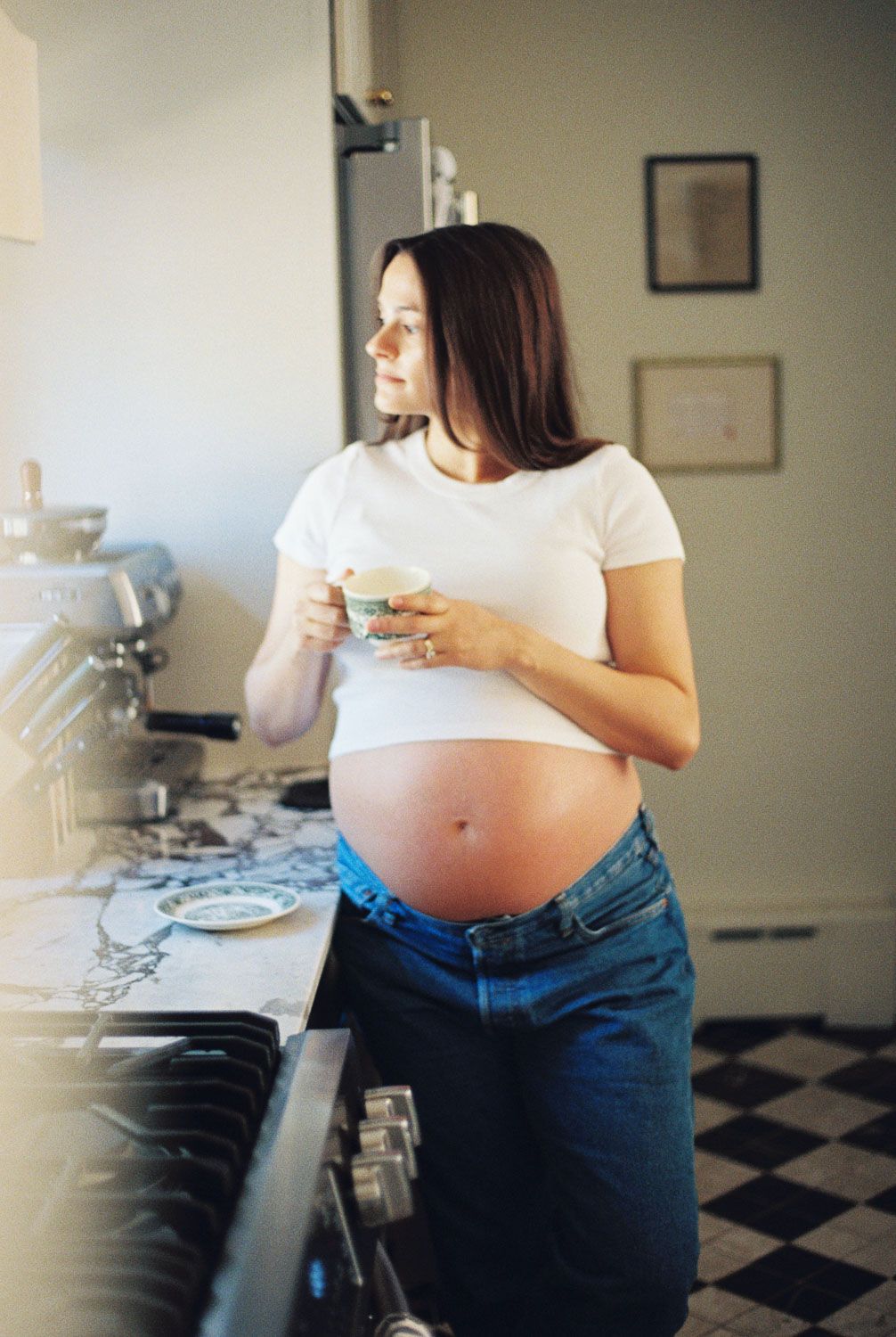 Pregnant woman in white tee and jeans holding a cup in a kitchen.