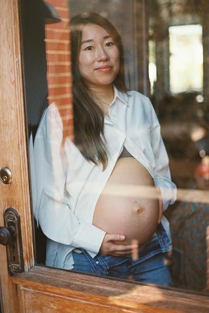 Pregnant Asian woman in open white shirt and jeans cradling baby bump, smiling through glass door.