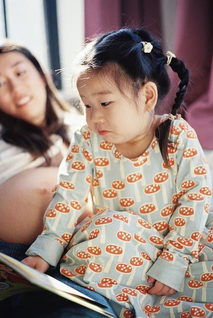 Young Asian girl in mushroom-print pajamas reads a book while her pregnant mother smiles warmly in the background.