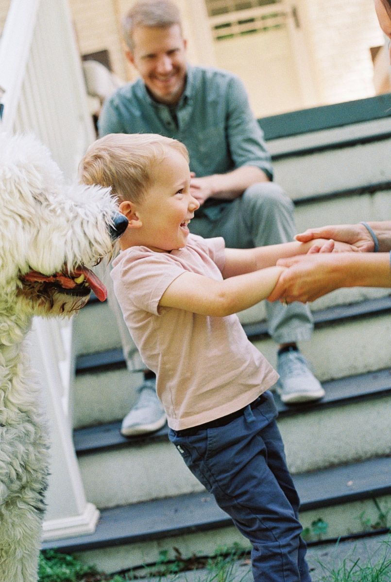 Happy toddler laughing while playing with a fluffy white dog on porch steps with father watching.