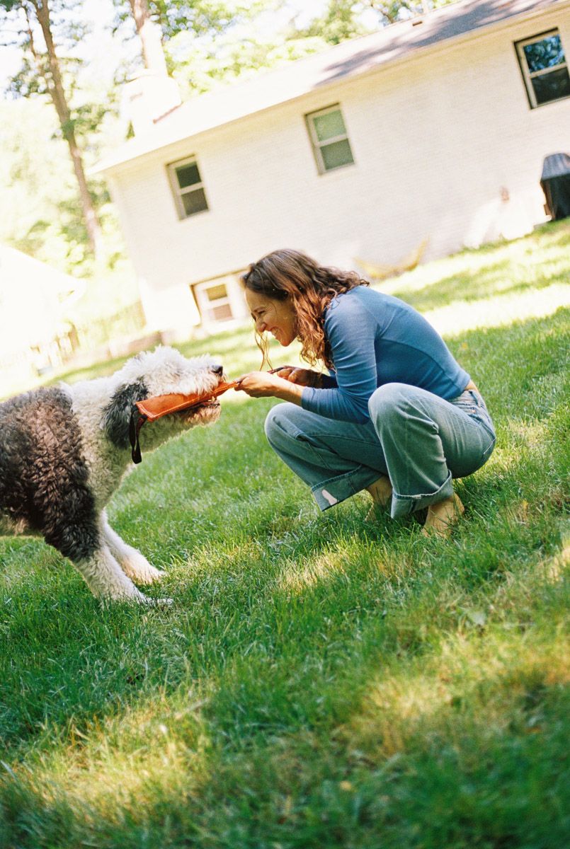 Woman in blue crouching on grass playing tug-of-war with a fluffy dog using an orange toy in a backyard.