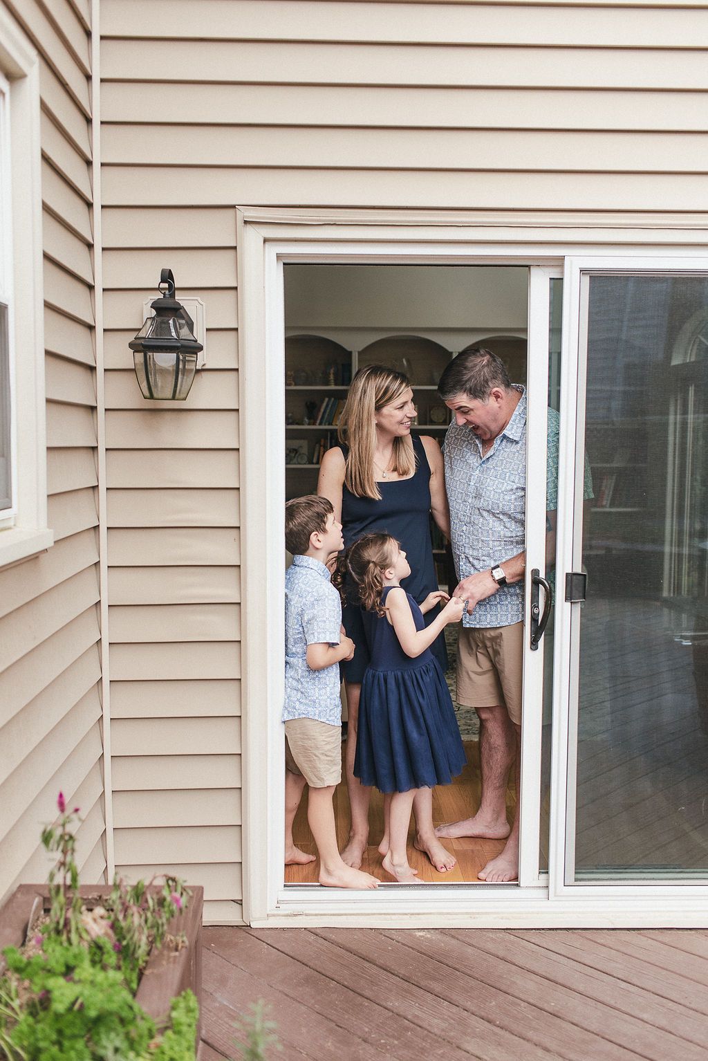 Family members interact playfully through a row of windows on a beige-sided house exterior.