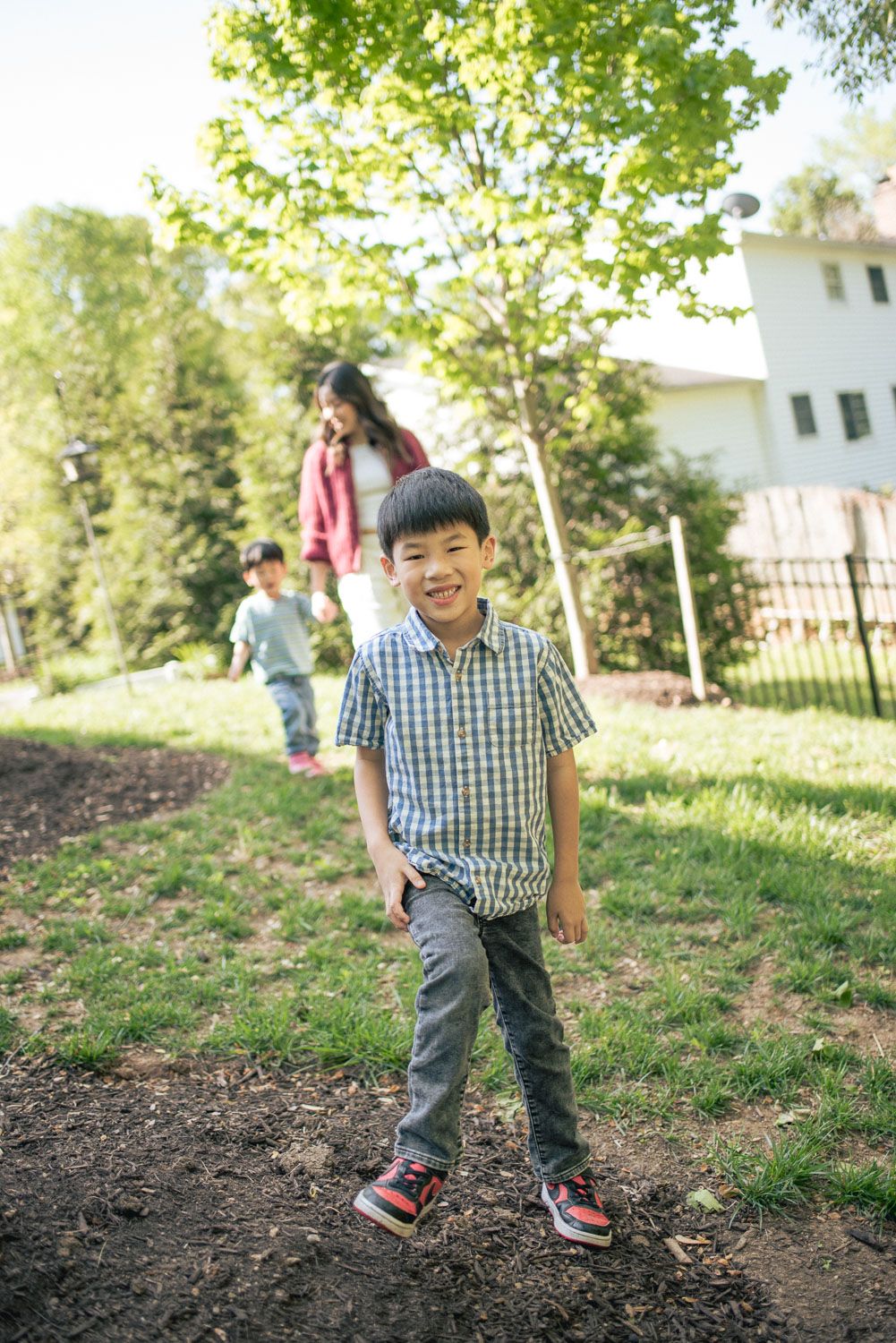Smiling Asian boy in plaid shirt walks outdoors in sunny backyard with woman and child in background.