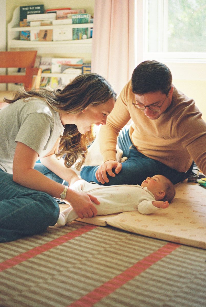 A smiling woman and man in casual clothes lean over a newborn baby lying on a padded play mat in a sunlit living room.