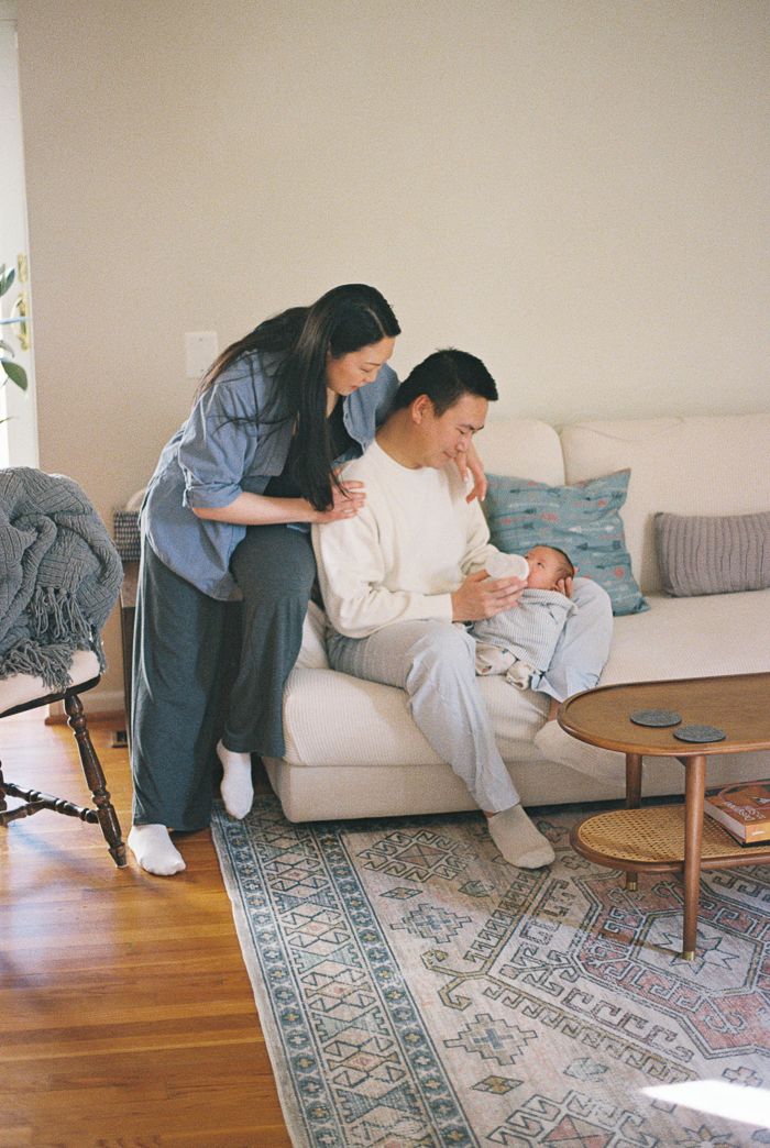 A couple admires their newborn baby on a white sofa in a cozy living room with hardwood floors.