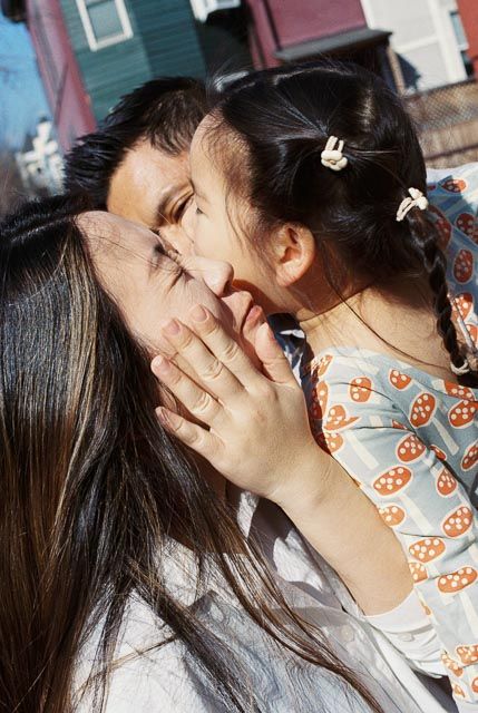 A young child whispers into her mother's ear while being held outdoors on a sunny day.
