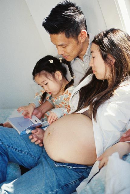Pregnant Asian woman and man reading a book with their young daughter, enjoying a family moment together.