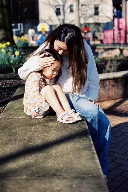A mother tenderly comforts her young daughter while sitting together on a stone ledge in a sunny park.