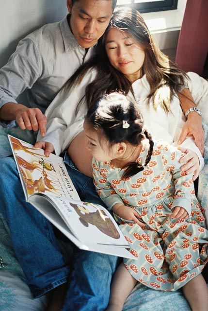 Asian family of three reading a colorful picture book together, with pregnant mother, father, and young girl in floral dress.