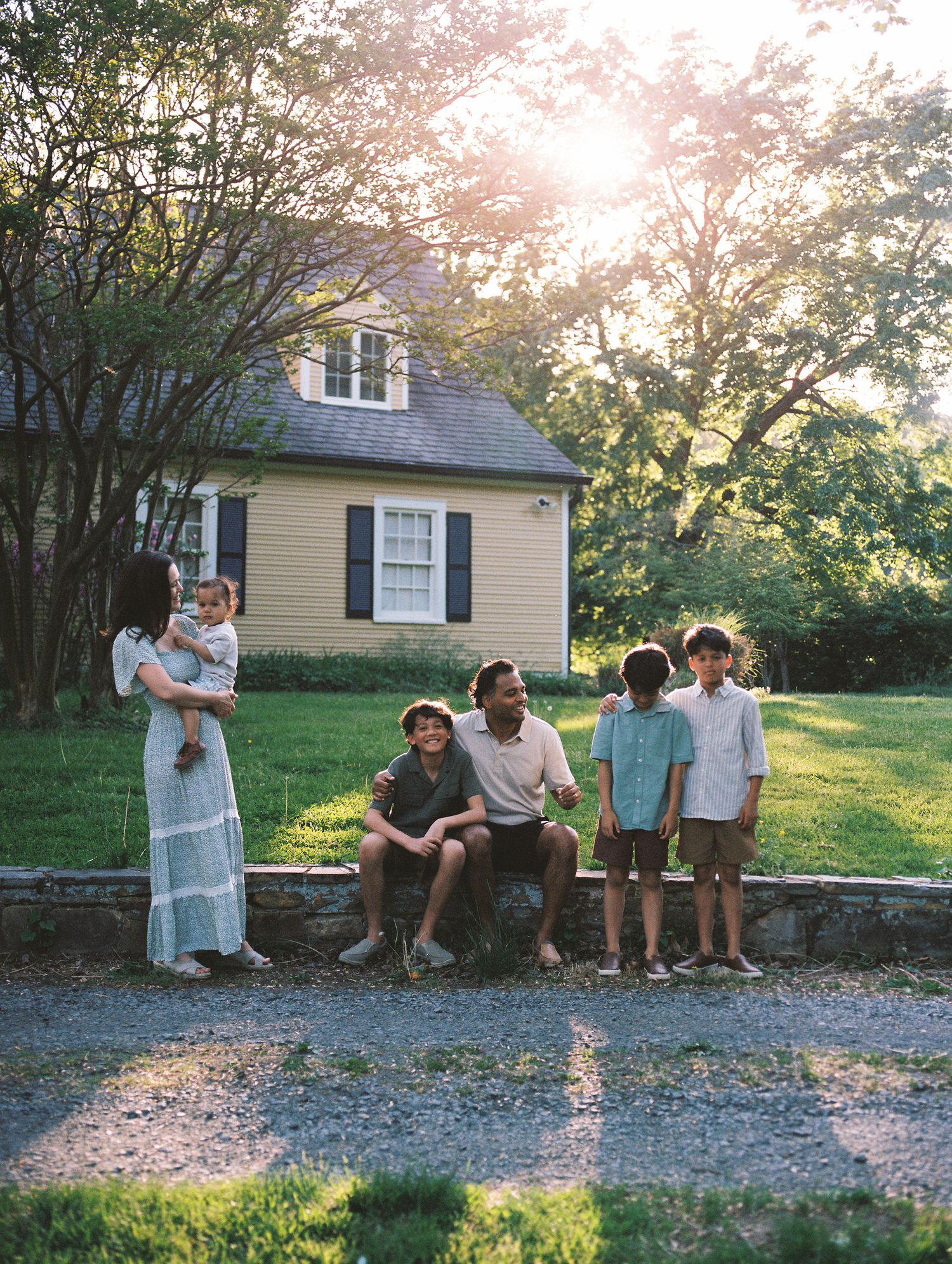 A family gathering outside a yellow house at sunset with children and adults sitting on a stone wall.