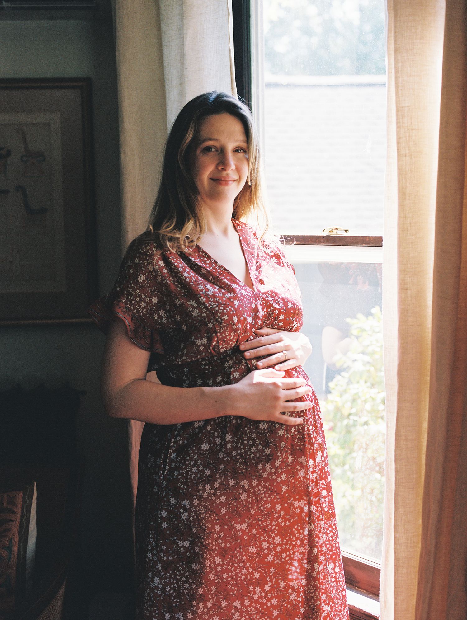 Expectant woman in a floral red dress stands by a sunlit window, gently holding her baby bump.