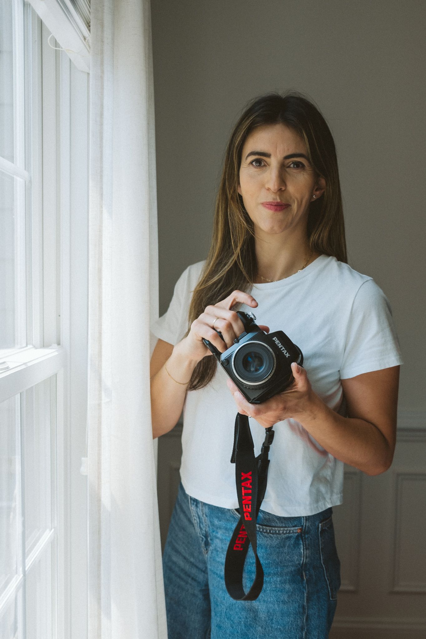 Photographer in white t-shirt and jeans stands by window holding professional camera with red strap.