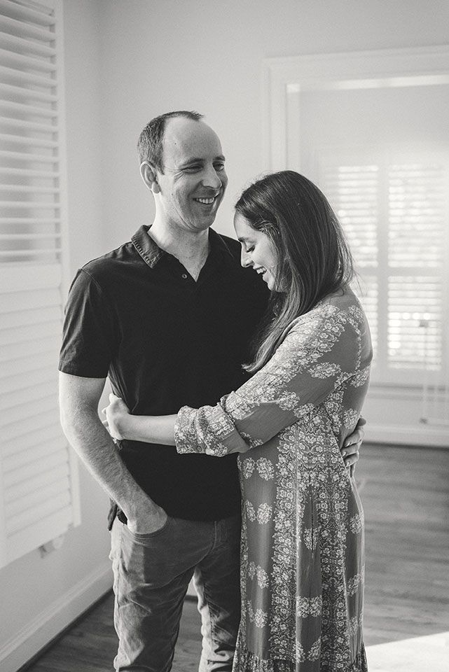 A couple shares an intimate black and white moment together in a sunlit room with window blinds.