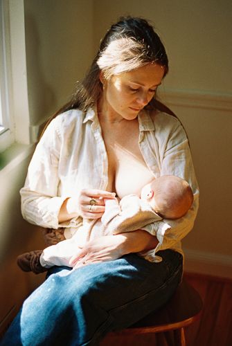 Mother breastfeeding newborn baby while seated by sunny window in white linen shirt and jeans.