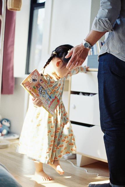 A young girl in a floral dress reads a book as an adult gently places a hand on her head indoors.
