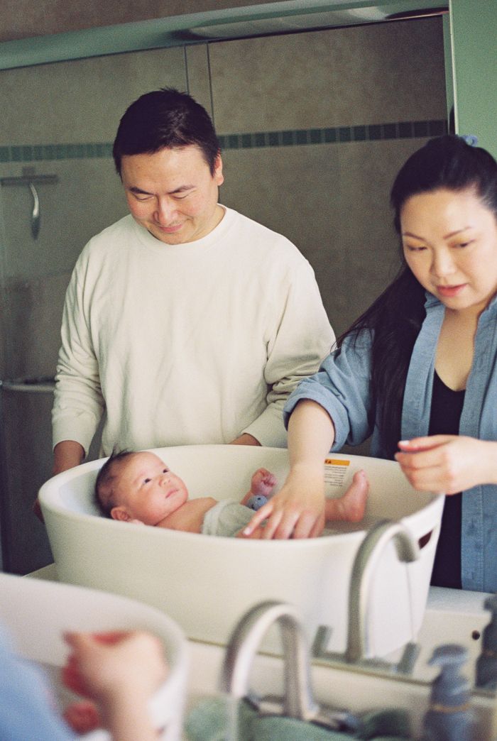 Parents smiling while bathing their newborn baby in a small white tub in a hospital room.