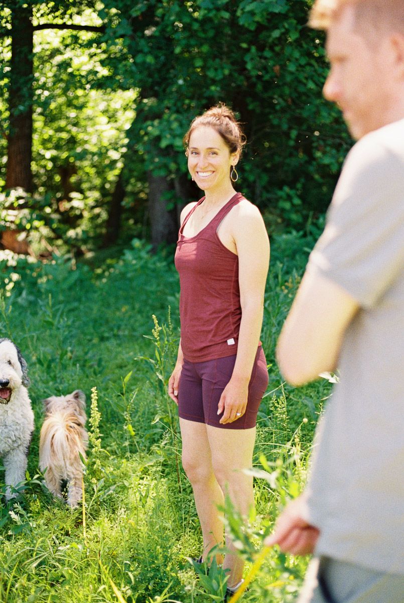 A smiling woman in a maroon tank top and shorts stands in a lush green forest with two dogs nearby.