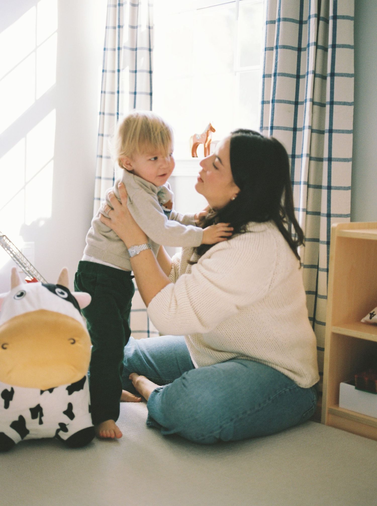 A parent and toddler share playful moments together while sitting on a floor cushion near a window.