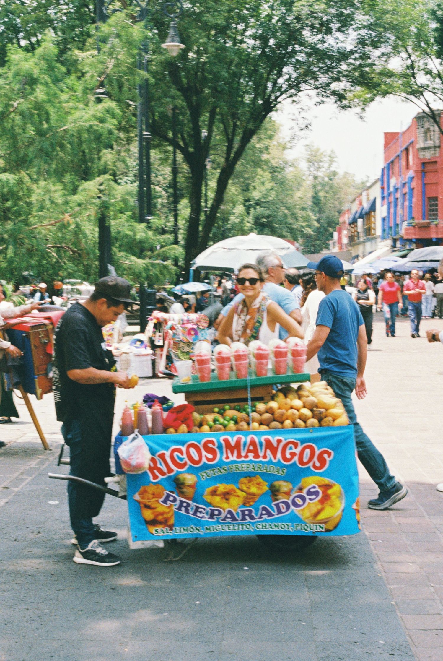 Street vendors prepare colorful Mexican snacks and treats at an outdoor market on a sunny day.