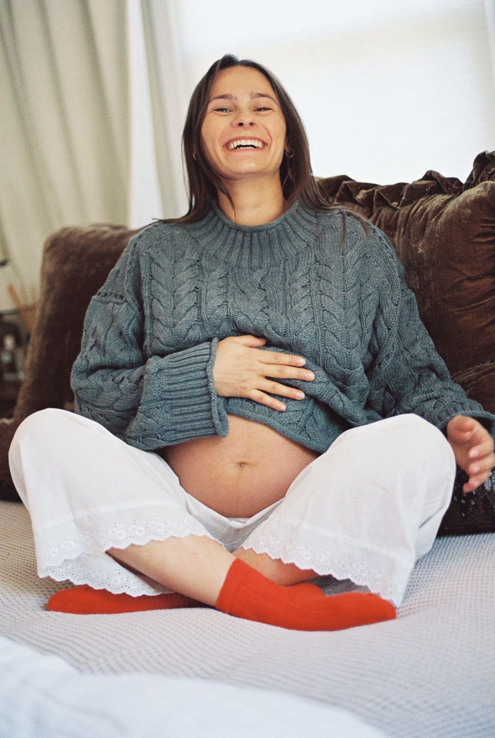 Laughing pregnant woman in gray cable-knit sweater and white pants sitting cross-legged on couch holding her belly.