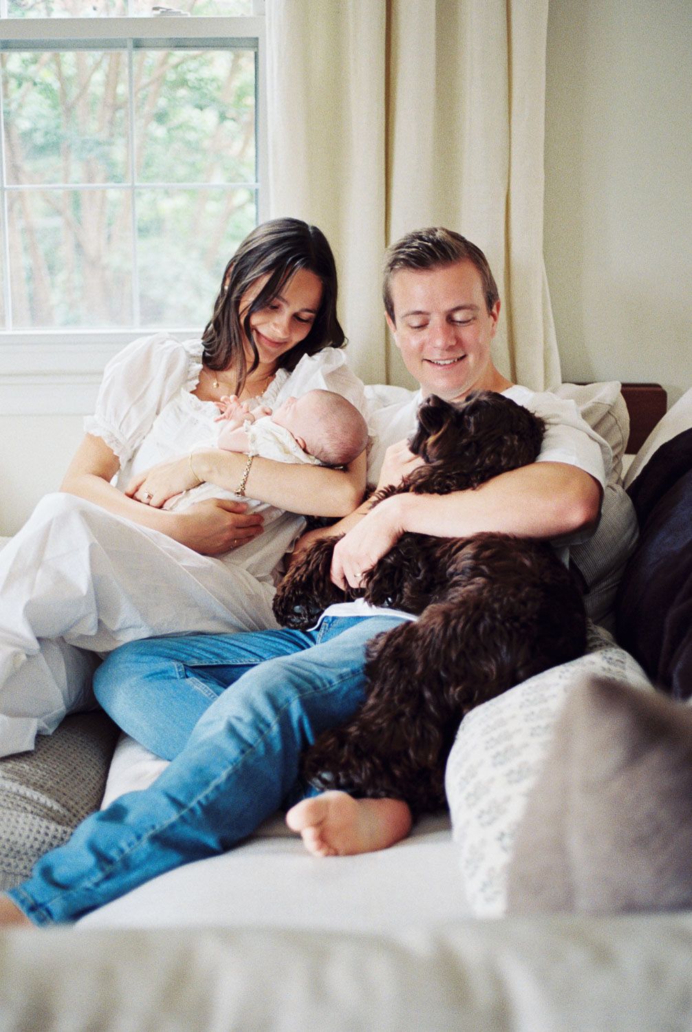 Young family with newborn baby and black dog relaxing together on cozy white couch near window.